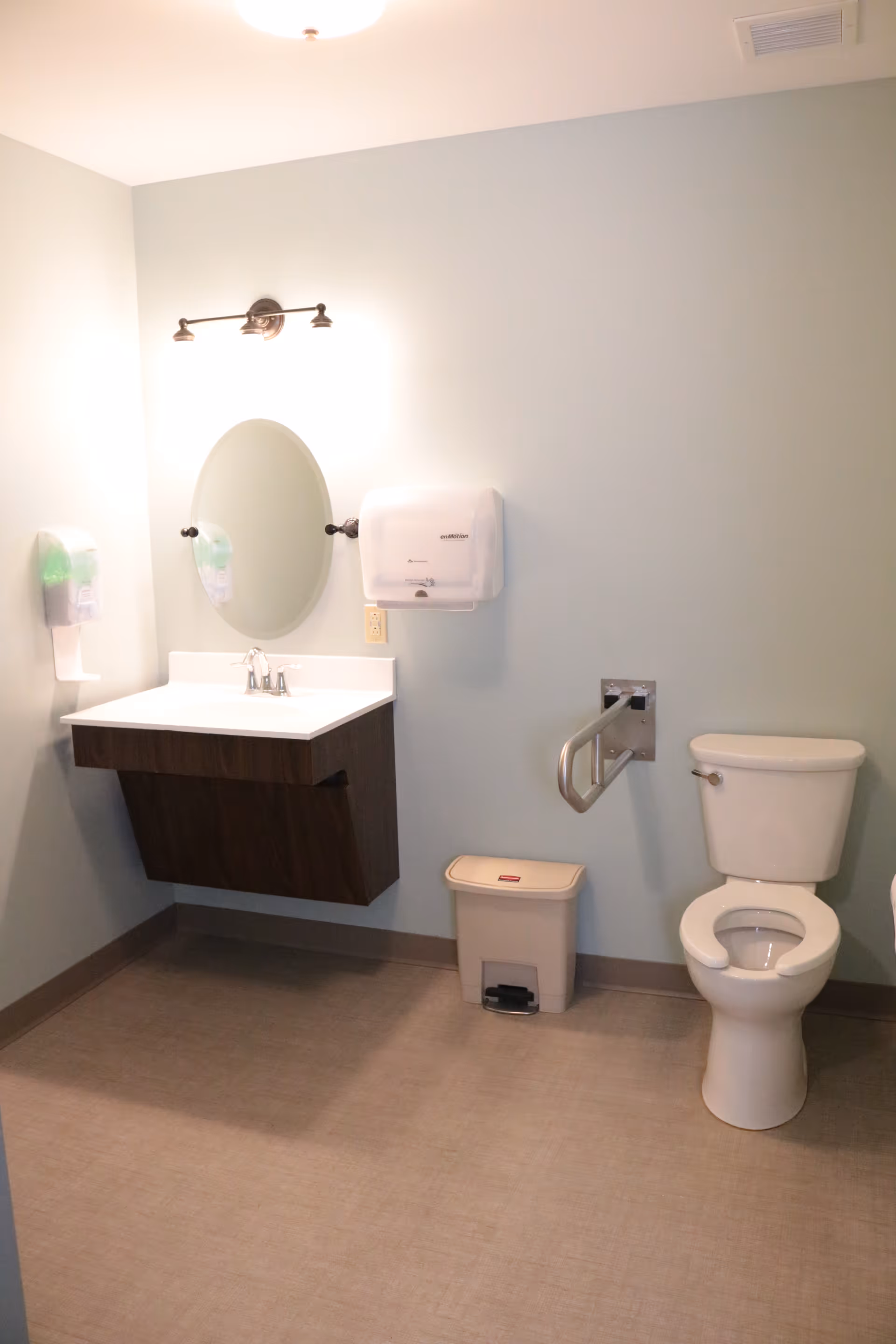 A clean and accessible bathroom with a white toilet, a wall-mounted sink with a dark wooden cabinet, an oval mirror above the sink, a wall-mounted soap dispenser, a paper towel dispenser, a small trash bin, and a grab bar next to the toilet.