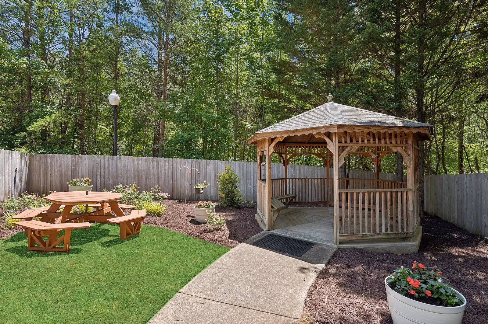 Outdoor area with a wooden gazebo on the right and a round wooden picnic table with attached benches on the left, surrounded by a green lawn, flower pots, and a wooden fence with trees in the background.