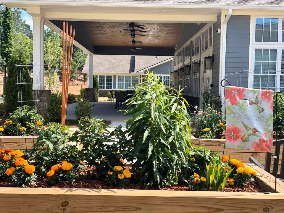 Outdoor garden area at Manor Lake Assisted Living & Memory Care - Gainesville featuring raised wooden garden beds with green plants and orange flowers. A colorful welcome flag with hummingbirds and flowers is displayed on the right side. The garden is situated under a covered patio area with ceiling fans and outdoor seating visible in the background.