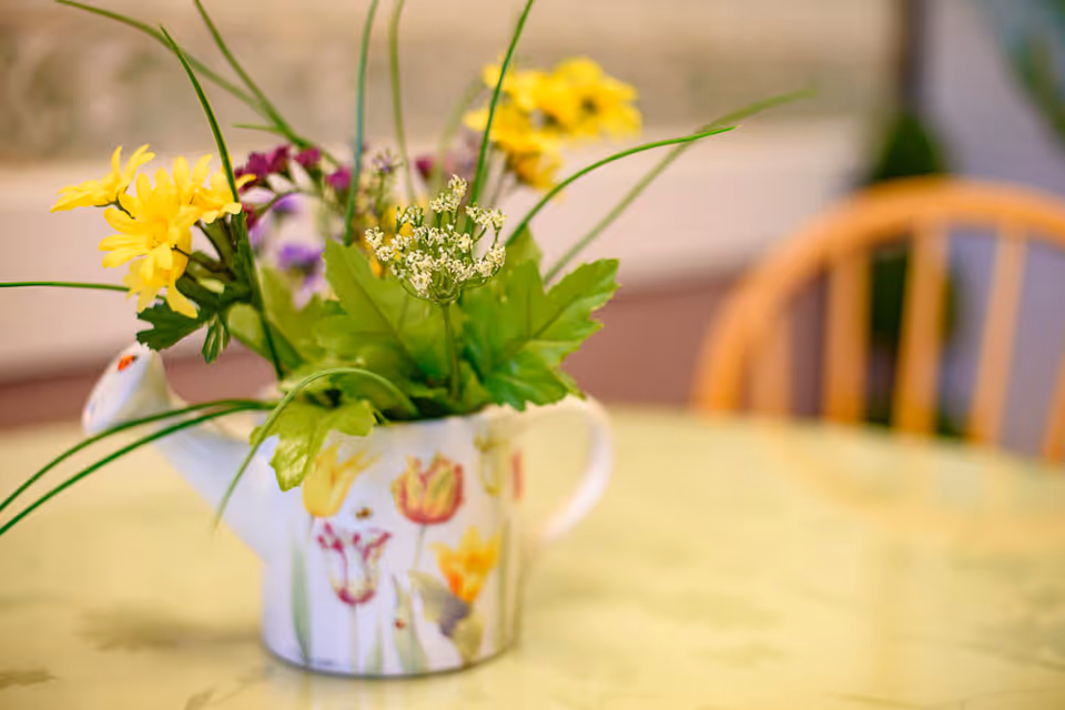 A floral arrangement in a decorative teapot sits on a table with a wooden chair blurred in the background.