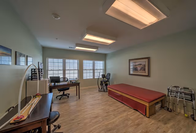 Sunlit therapy room with a red treatment table, desk, exercise bike, and walkers on wood flooring.