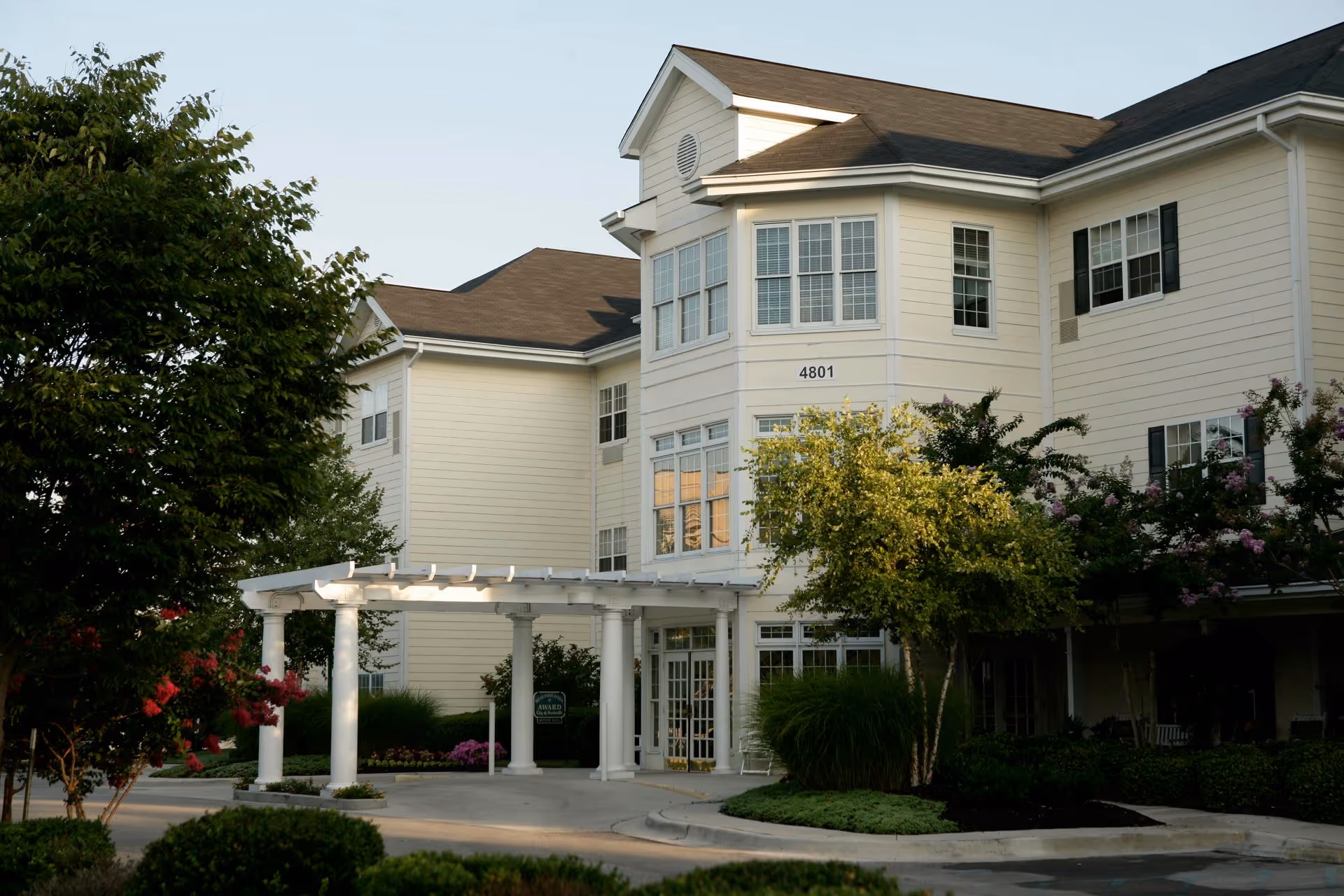 Exterior view of a multi-story assisted living facility building with beige siding, multiple windows, and a covered entrance supported by white columns. The building is surrounded by landscaped greenery including trees, bushes, and flowering plants.