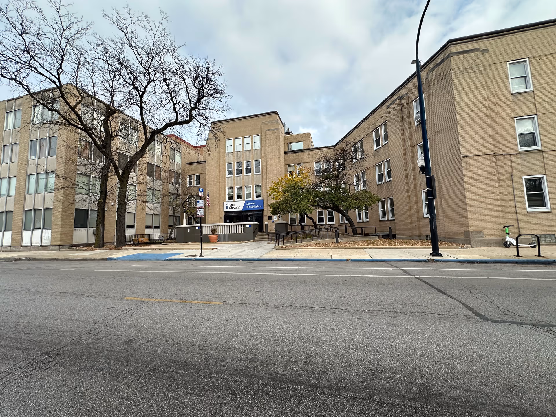 Exterior front view of a multi-story brick rehabilitation center building with bare trees, a sidewalk and a street in the foreground.