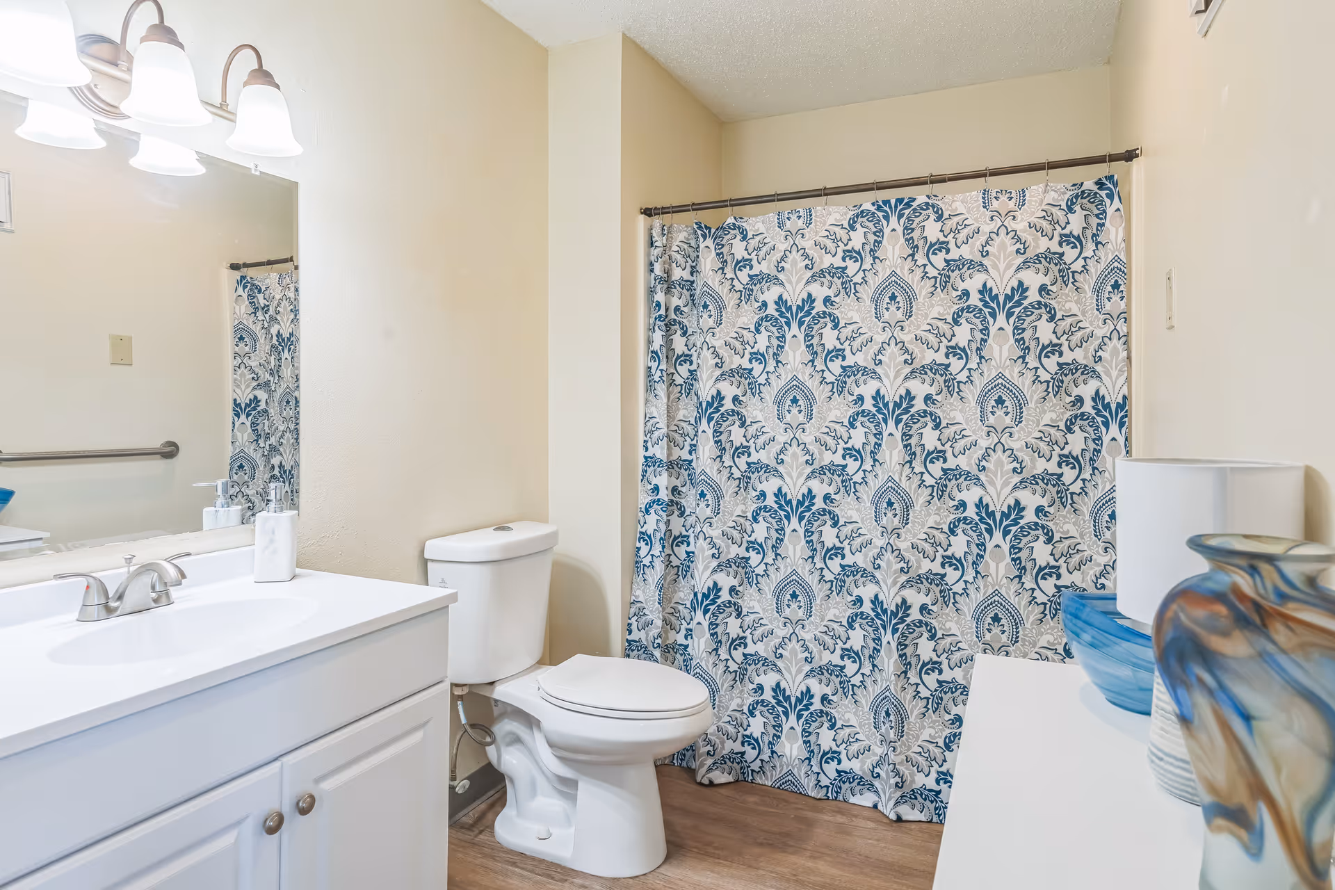 A clean bathroom with a white vanity and sink, a large mirror with three light fixtures above it, a white toilet, and a shower with a blue and white patterned curtain. There is a white cabinet on the right side with decorative vases and a lamp on top. The floor is wood-style.