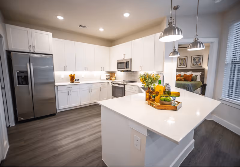 Modern kitchen with white cabinets, stainless steel refrigerator, oven, and microwave. A large white island countertop is in the center with decorative items including a tray with drinks and glasses. Pendant lights hang above the island. A bedroom with a bed and framed artwork is visible through an open doorway.