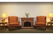 Two upholstered armchairs flank a small decorative brick fireplace with matching side tables and lamps in a sitting area.