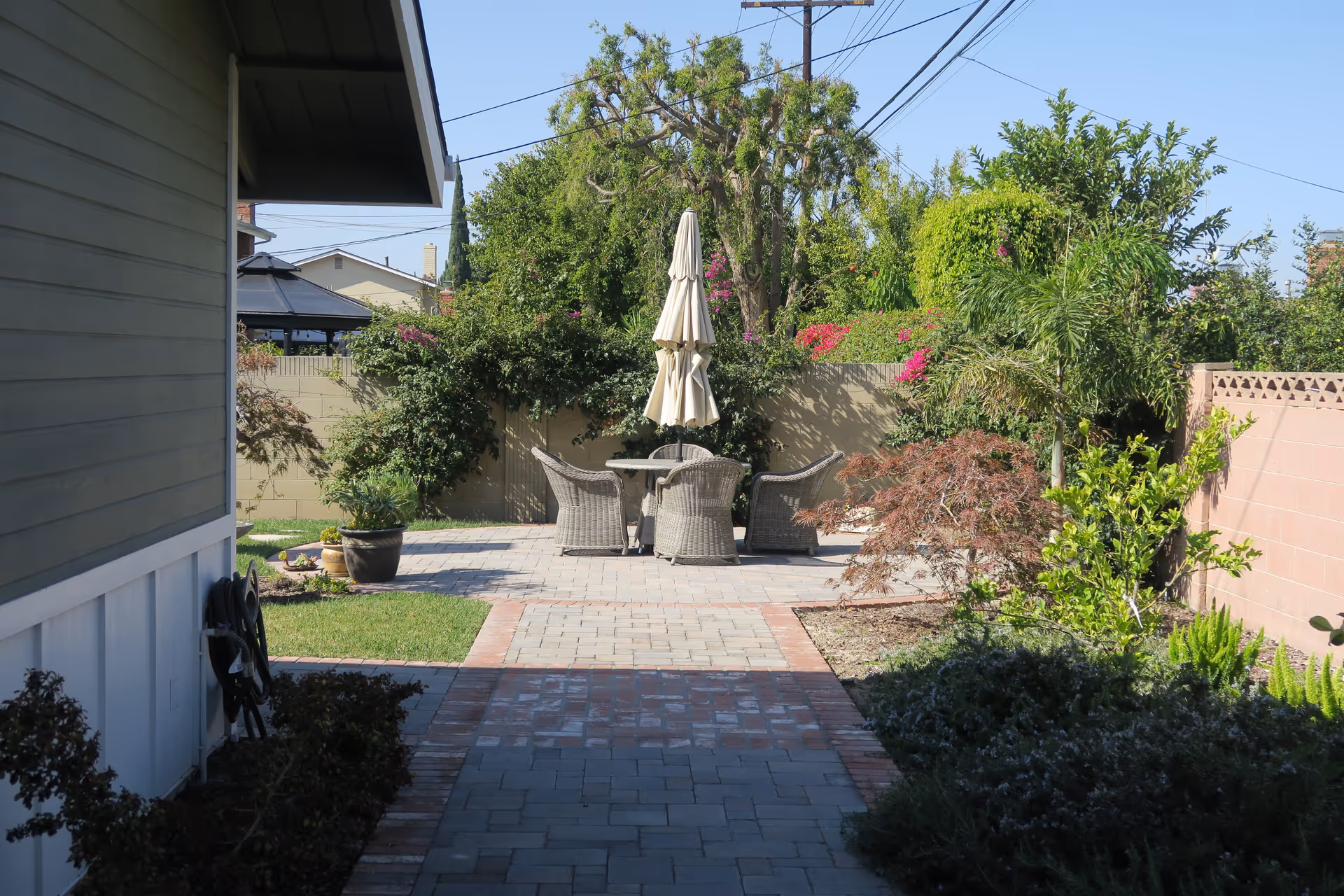 Backyard patio with a round table, four wicker chairs and a closed umbrella surrounded by garden beds and a paved walkway.