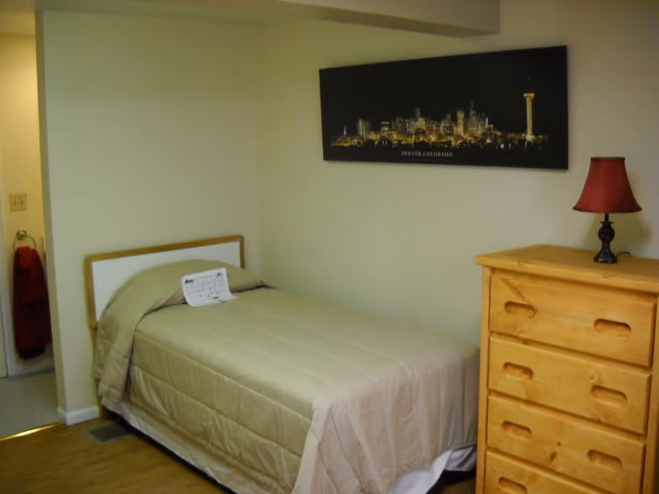 A small bedroom with a single bed covered in a beige comforter. There is a wooden dresser with a red lamp on top to the right of the bed. A framed picture of the Denver, Colorado skyline hangs on the wall above the dresser. A doorway to the left leads to another room, with a red towel hanging on a ring near the door.