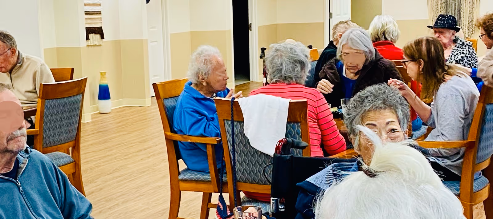 Several elderly residents sit and converse at dining tables in a communal dining/activity room.