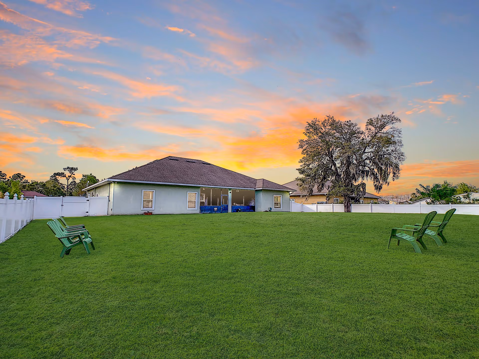 A large grassy backyard area with four green plastic chairs arranged in two pairs facing each other. In the background, there is a single-story building with a sloped roof and several windows. A large tree stands to the right side of the building. The sky is colorful with a sunset featuring orange and pink clouds.