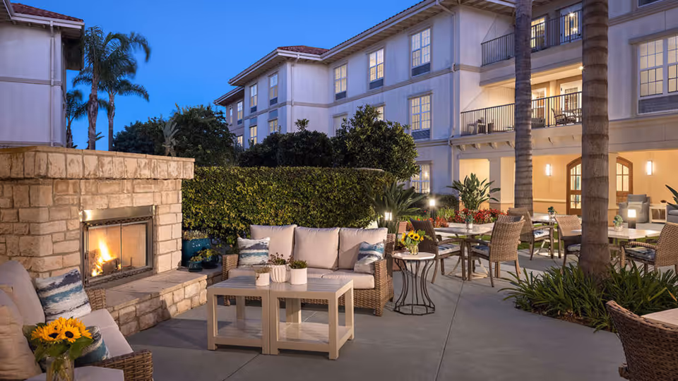 Outdoor patio area at dusk with wicker seating, tables, a stone fireplace with a fire burning, potted plants, and a multi-story building in the background with lit windows.