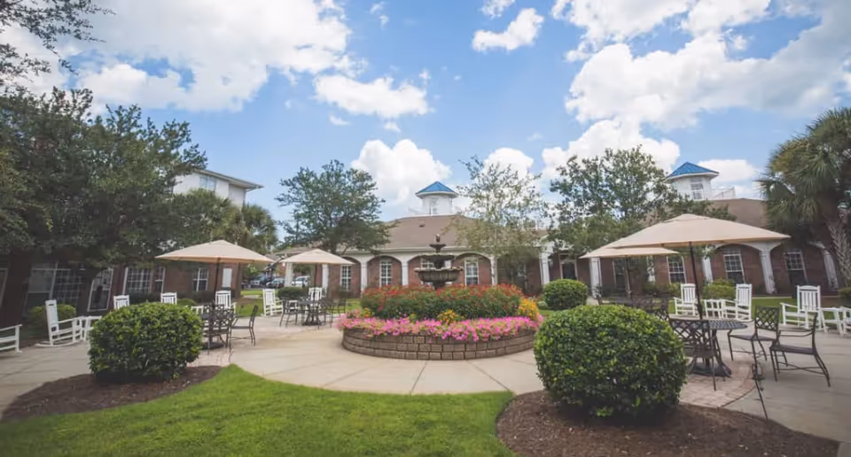Outdoor courtyard area at The Lakes at Litchfield featuring a circular flower bed with a fountain in the center, surrounded by patio tables with umbrellas and chairs. The area is landscaped with bushes, trees, and grass, with a brick building in the background under a partly cloudy sky.