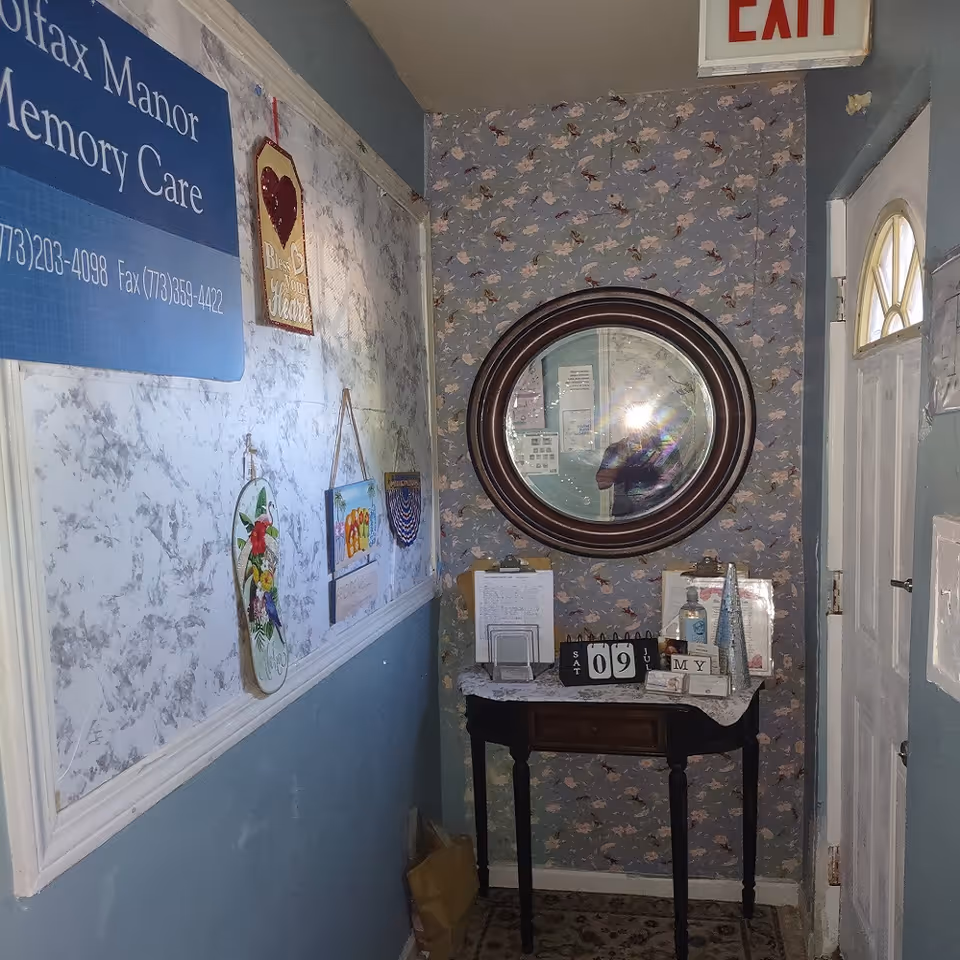 A small hallway corner in Colfax Manor Assisted Living with a round mirror on floral wallpapered wall above a small table holding clipboards, a calendar showing Saturday July 9, hand sanitizer, and decorative items. The adjacent wall has a blue sign with contact information and several hanging decorative plaques. An exit sign is visible above a white door with a small window.