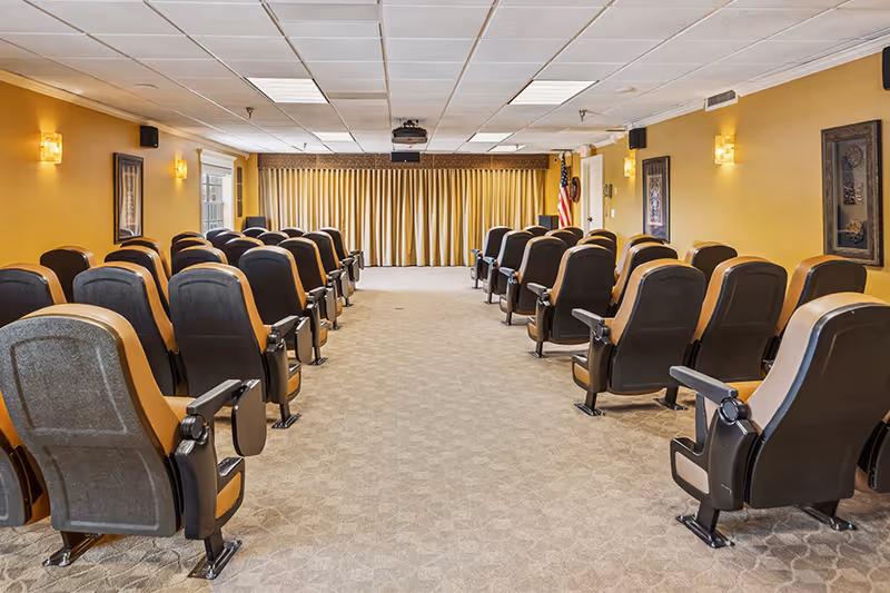 Room with rows of theater-style seating facing a stage area with closed gold curtains, beige carpet, yellow walls, framed artwork, and an American flag in the corner.