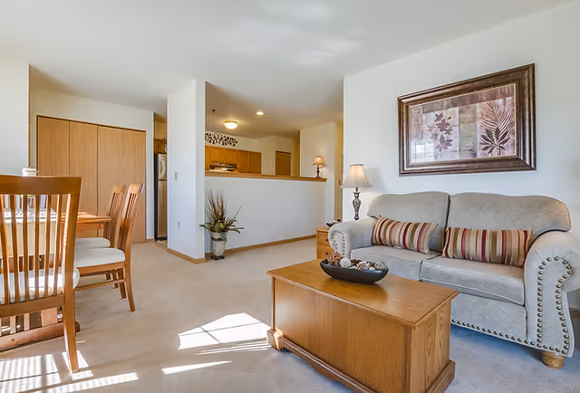 A bright and cozy living room and dining area in a senior living facility. The living room features a beige sofa with striped cushions, a wooden coffee table with decorative items, a side table with a lamp, and a framed floral artwork on the wall. The dining area has a wooden dining table with matching chairs. The kitchen is partially visible behind a half wall, with wooden cabinets and a refrigerator. The room is carpeted and well-lit with natural light coming through a window.