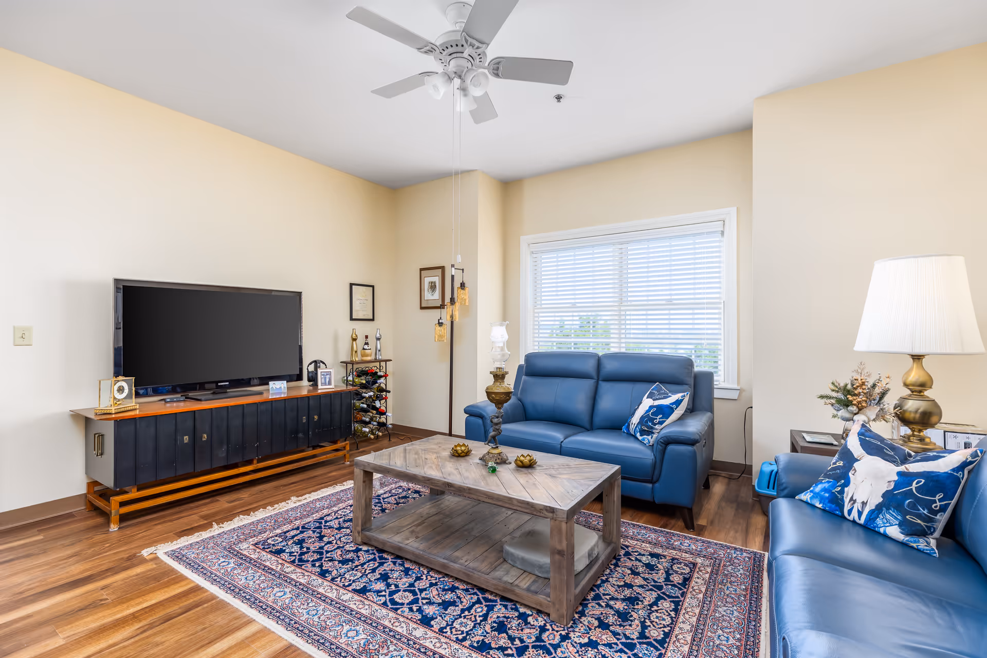 A bright living room with beige walls and wooden flooring featuring two blue leather sofas adorned with decorative pillows. A wooden coffee table sits on a patterned blue and red rug in the center. A large flat-screen TV is placed on a black and wood TV stand against one wall. A window with white blinds lets in natural light, and a white ceiling fan is mounted above. There is a floor lamp with three hanging lights and a table lamp with a white shade on a side table next to one of the sofas.