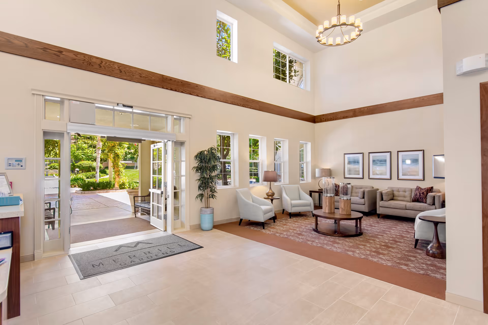 Bright and spacious senior living facility lobby with high ceilings, large windows, and an open entrance door leading outside. The seating area includes two armchairs, two sofas, a round coffee table with decorative items, floor lamps, and framed artwork on the wall. A potted plant is placed near the windows.