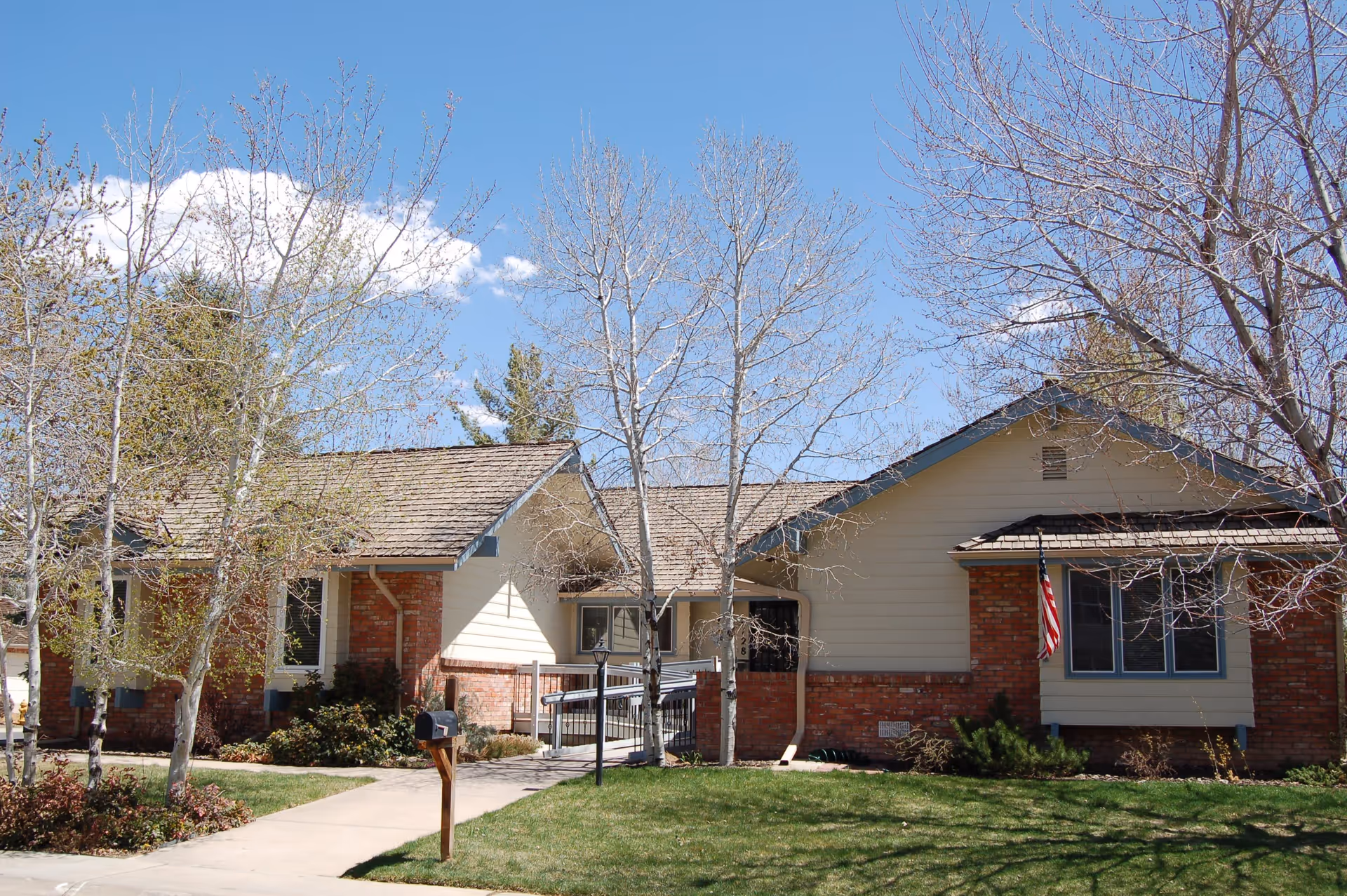 Exterior view of a single-story residential building with a brick and beige siding facade, a shingled roof, several leafless trees in the front yard, a concrete walkway leading to the entrance, and an American flag mounted near a window.