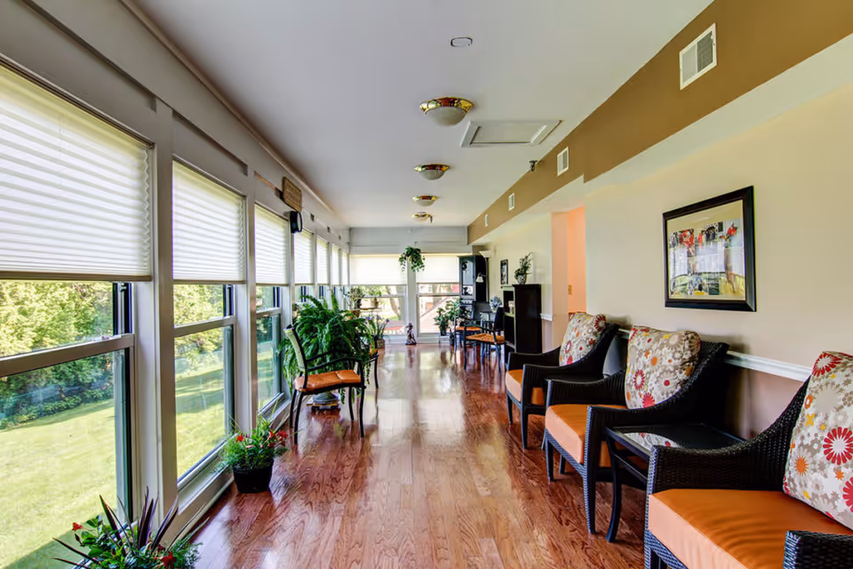 A bright and spacious hallway in a senior living community with large windows on one side letting in natural light. The hallway features wooden flooring, several cushioned chairs with floral patterned pillows along the opposite wall, and various potted plants placed near the windows and at the end of the hallway. The walls are painted in neutral tones with framed artwork hanging on one side.