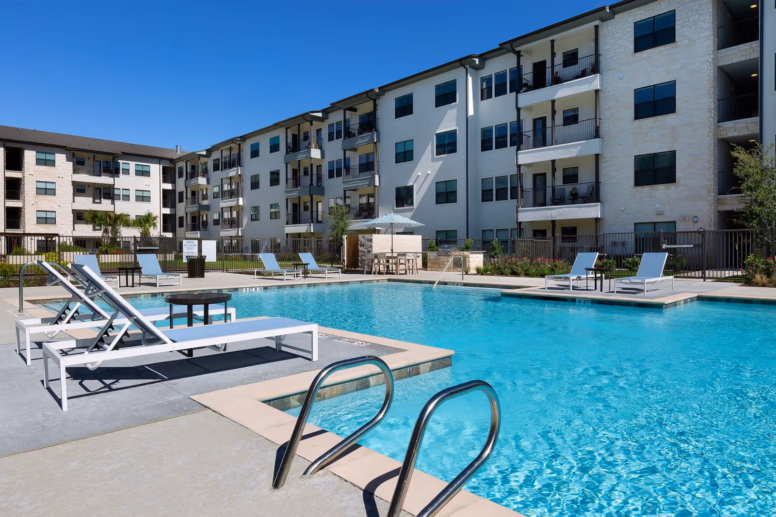 Outdoor swimming pool with lounge chairs in front of a multi-story residential building under a clear blue sky.