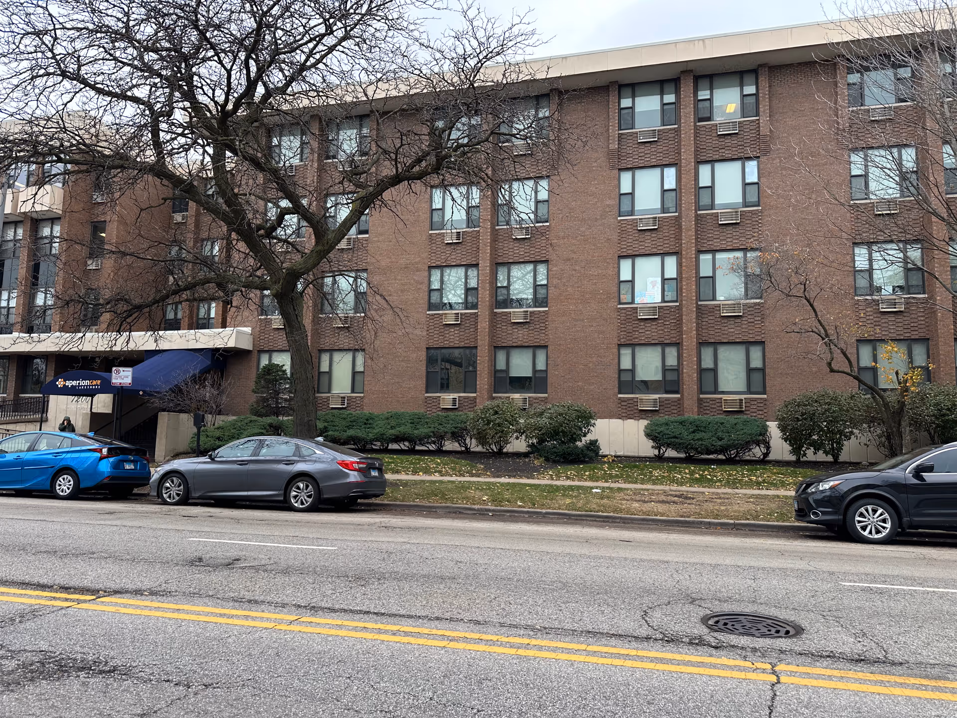 Exterior view of a multi-story brick building with several windows, a blue awning entrance labeled 'aperioncare Lakeshore,' a few parked cars along the street, and leafless trees in front of the building.