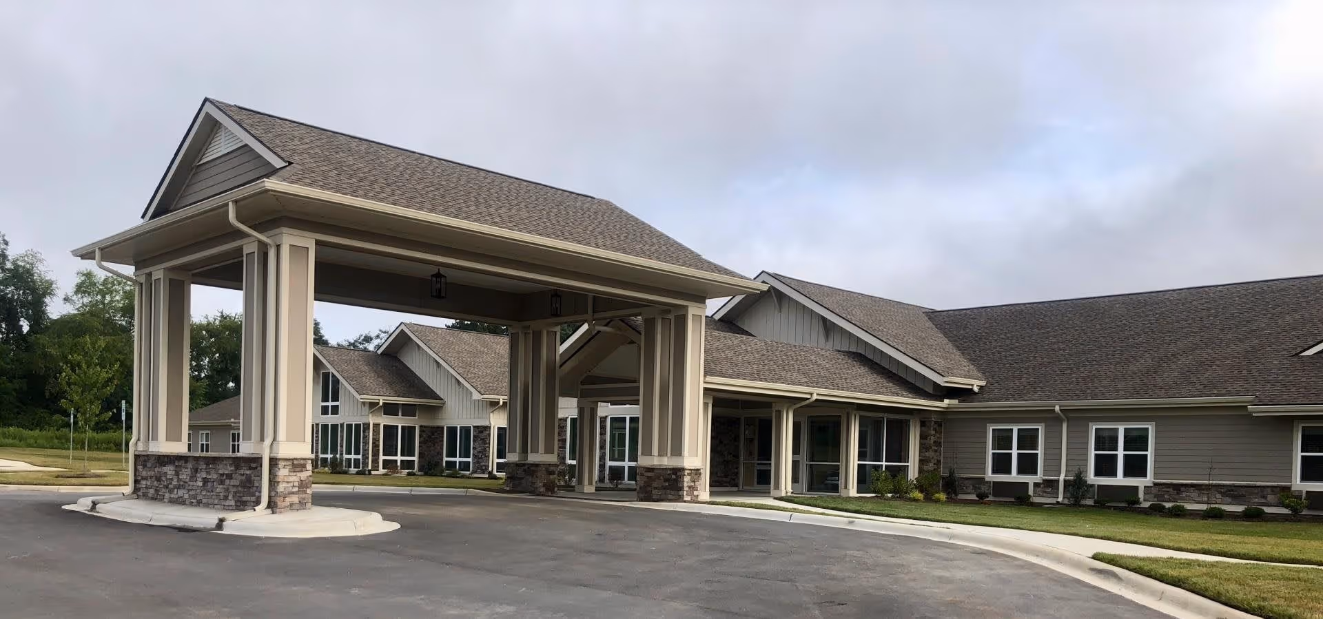 Exterior view of The Landings of Mills River senior living facility showing a covered entrance with stone and beige pillars, multiple windows, and a well-maintained lawn under a cloudy sky.