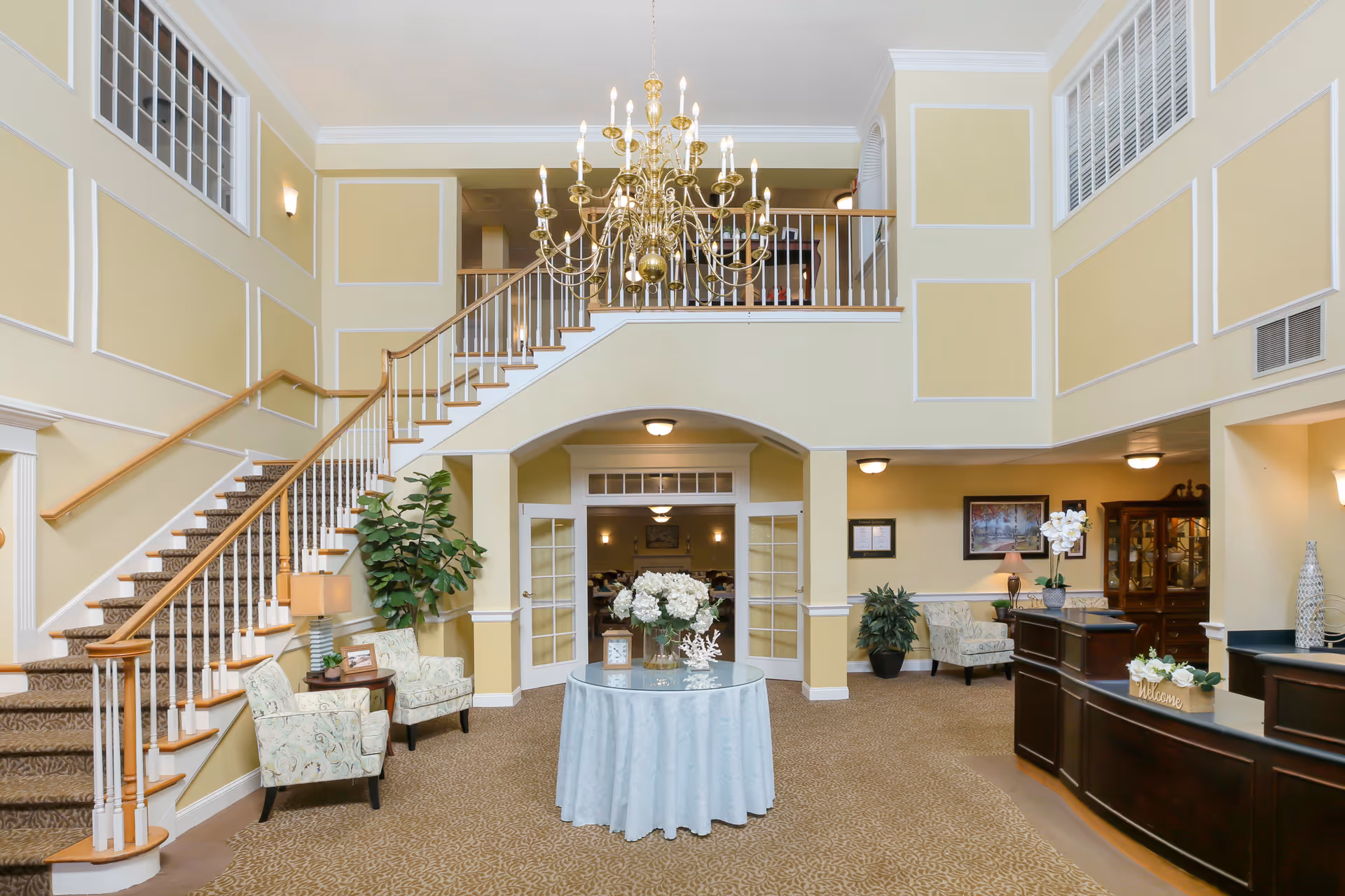 A spacious and elegant senior living facility lobby with a large chandelier hanging from the ceiling. There is a staircase with wooden handrails on the left, two upholstered chairs with a small table and lamp beside them, and a round table with a floral centerpiece in the middle. The reception desk is on the right side with decorative items and a 'Welcome' sign. The walls are painted light yellow with white trim, and there are plants and framed pictures adding to the decor.