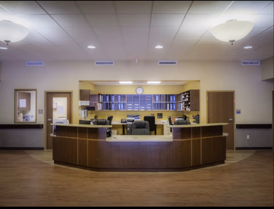 Reception desk area in a senior living facility with a curved wooden counter, office chairs, filing cabinets, and shelves with blue binders. The room has beige walls, wooden doors, and ceiling lights.