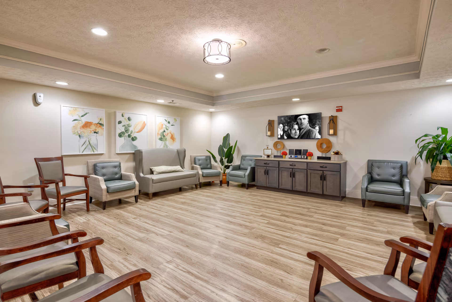 A spacious, well-lit living room with wooden flooring, multiple chairs arranged around the perimeter, a gray loveseat, and armchairs. The walls are decorated with three floral paintings and a mounted flat-screen TV displaying a black and white movie scene. There are plants and decorative items on a long cabinet beneath the TV.
