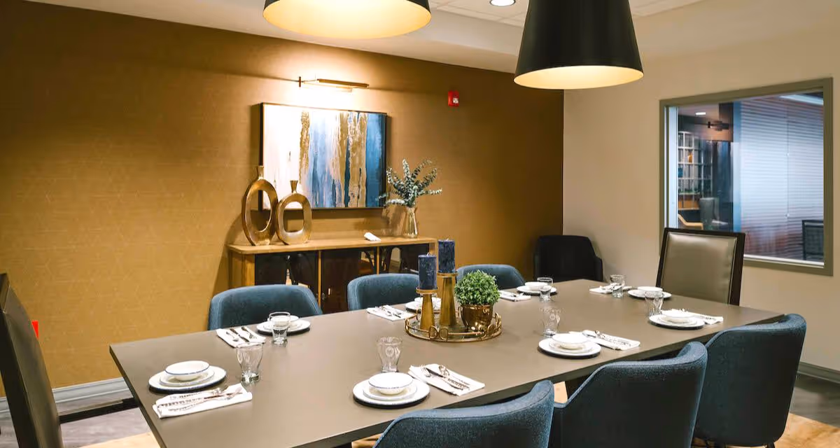 Modern dining room with a long table set with plates and glassware, blue upholstered chairs, pendant lights, and a decorative sideboard.