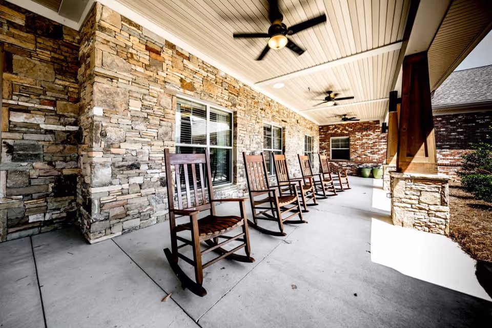 Covered outdoor patio area with a row of wooden rocking chairs lined up against a stone and brick wall. The ceiling is white with multiple ceiling fans, and there are large windows along the wall. The patio floor is concrete, and there are some plants visible at the far end.
