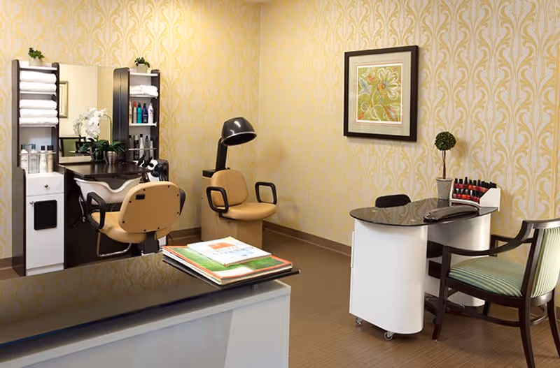 A well-lit salon room with a hair washing station, two beige salon chairs, a hair dryer, and a manicure table with nail polish bottles. The walls have a yellow patterned wallpaper, and there is a framed artwork hanging on the wall. The floor is a light brown wood or laminate.
