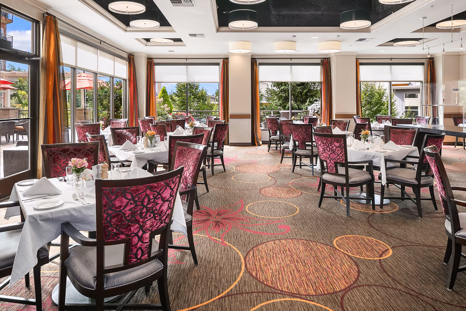 A bright and elegant dining room with multiple tables covered in white tablecloths, set with plates, glasses, and napkins. The chairs have dark wood frames with purple patterned upholstery. Large windows with orange curtains let in natural light and offer views of greenery and outdoor umbrellas. The carpet has a circular and floral pattern in shades of brown, orange, and red. Modern ceiling lights illuminate the room.