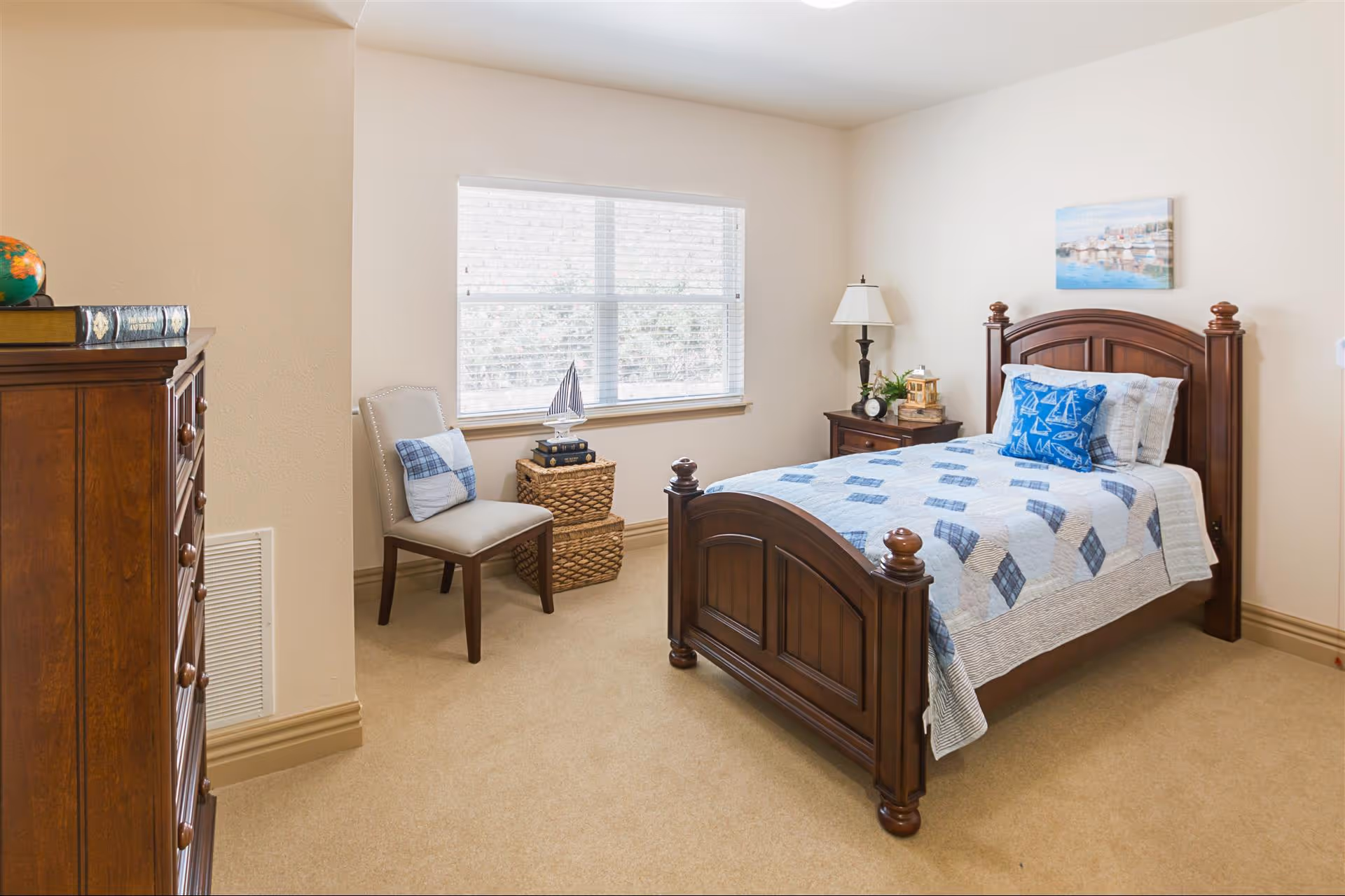 A cozy bedroom with a wooden bed frame featuring a blue and white quilt and matching pillows. Next to the bed is a wooden nightstand with a lamp, clock, and decorative items. A chair with a cushion is placed near a window with blinds, and two woven baskets are stacked beside it. A wooden dresser with books and a small globe is visible on the left side of the room. The walls are light-colored, and a painting hangs above the bed.