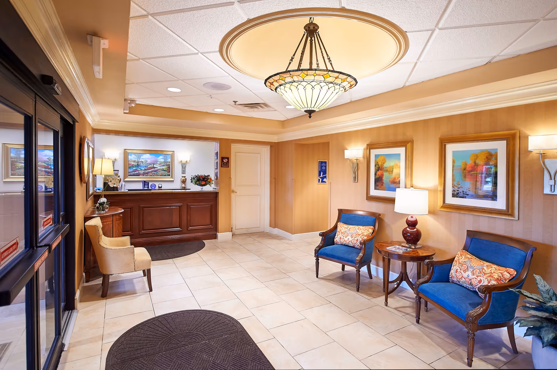 A bright and welcoming senior living facility lobby with beige tiled floors, warm beige walls, and a decorative ceiling light fixture. The space features a wooden reception desk with a lamp, flowers, and framed artwork behind it. There are two blue upholstered chairs with patterned cushions and a small round wooden table with a lamp between them. Additional framed paintings and wall sconces decorate the walls. A beige chair is positioned near the entrance door with glass panels.