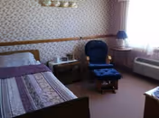 A senior living bedroom with a single bed covered in a patterned quilt, a wooden nightstand with framed photos and a lamp, a blue cushioned rocking chair with matching footrest, a window with curtains, and an air conditioning unit below the window.