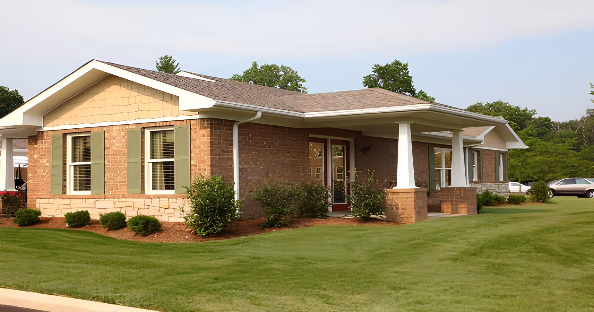 Exterior view of a single-story brick building with green shutters and a covered porch supported by white columns, surrounded by well-maintained grass and shrubs under a clear sky.