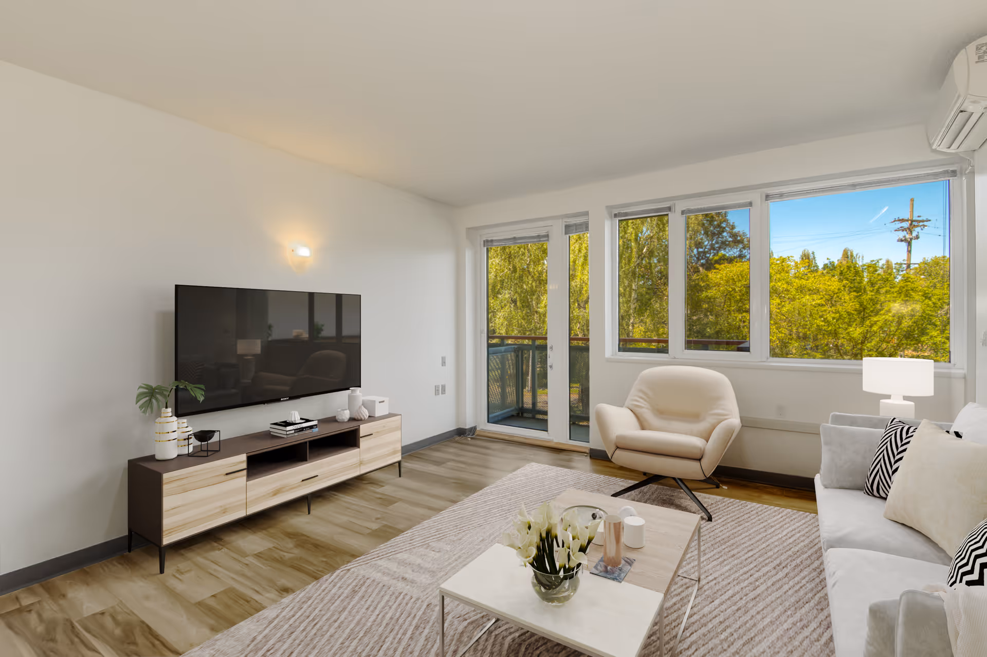 A bright living room with a large window and glass door leading to a balcony. The room features a modern TV on a wooden media console, a beige armchair, a white sofa with patterned pillows, a coffee table with a flower vase, and light wood flooring.