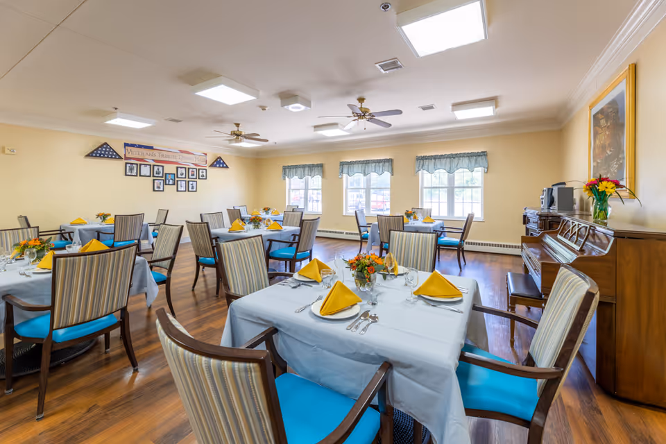 A bright dining room with several tables covered in light blue tablecloths, each set with yellow folded napkins, glassware, and silverware. The chairs have striped upholstery with blue seats. The room has wooden floors, three large windows with blue valances, ceiling fans, and a piano with a vase of flowers on top. A sign on the wall reads 'Veterans Tribute Dining Hall' with framed photos beneath it.