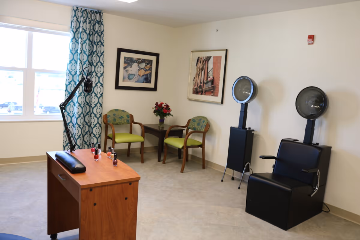 Salon-style room with two hooded hairdryers, a manicure table with nail polish bottles, and guest chairs by a window.
