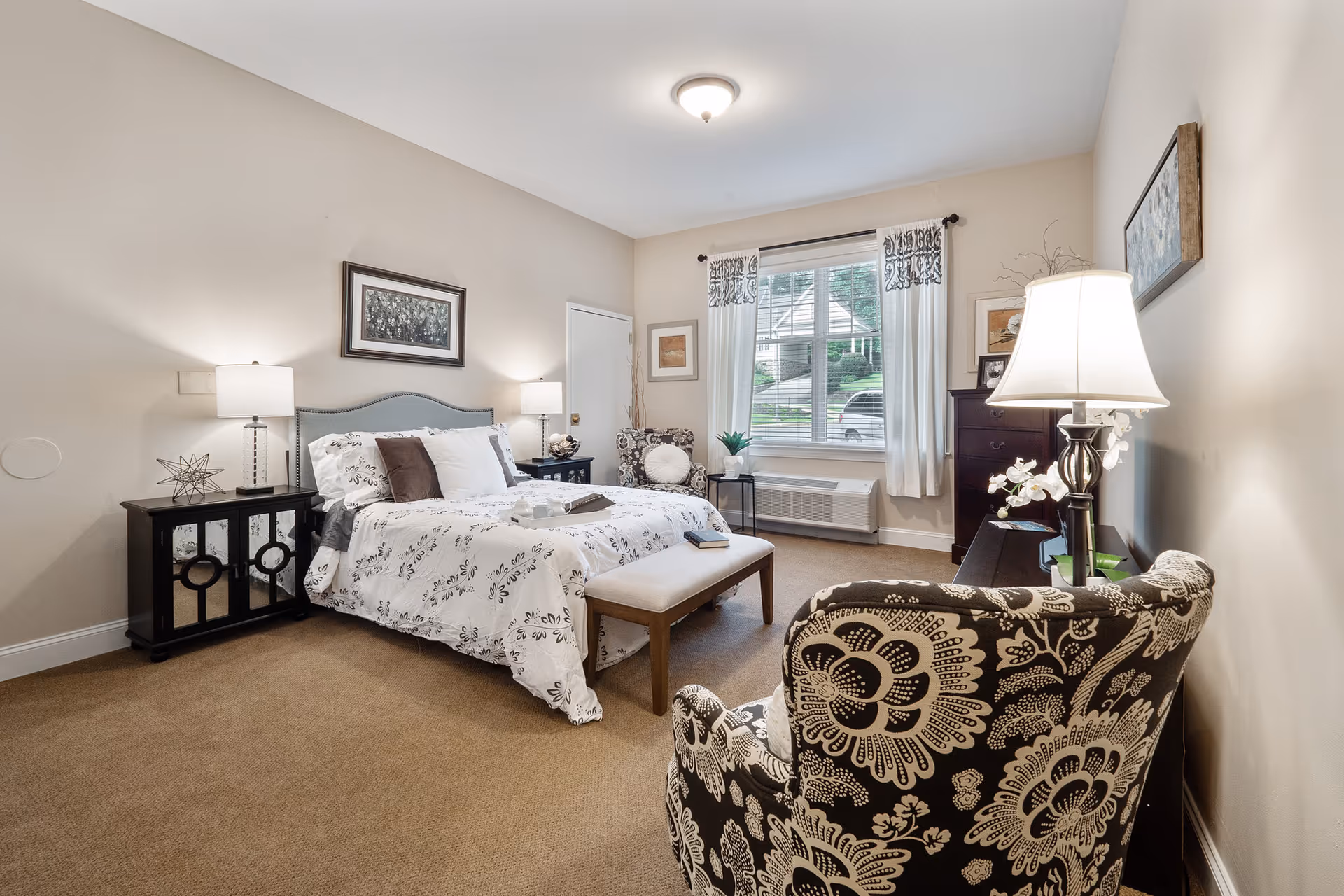 A well-lit bedroom with a large bed featuring white and floral bedding, two bedside tables with lamps, a cushioned bench at the foot of the bed, a patterned armchair, a window with white curtains, and a dresser with a lamp and decorative items.