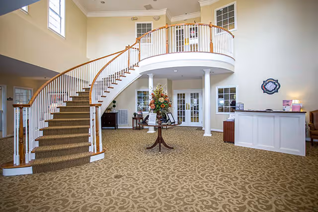 Spacious interior lobby area of a senior living facility with a curved staircase featuring wooden handrails and white balusters. The room has beige patterned carpet, cream-colored walls, and large windows letting in natural light. A round table with a floral arrangement is centered in the room, and a white reception desk is visible on the right side.