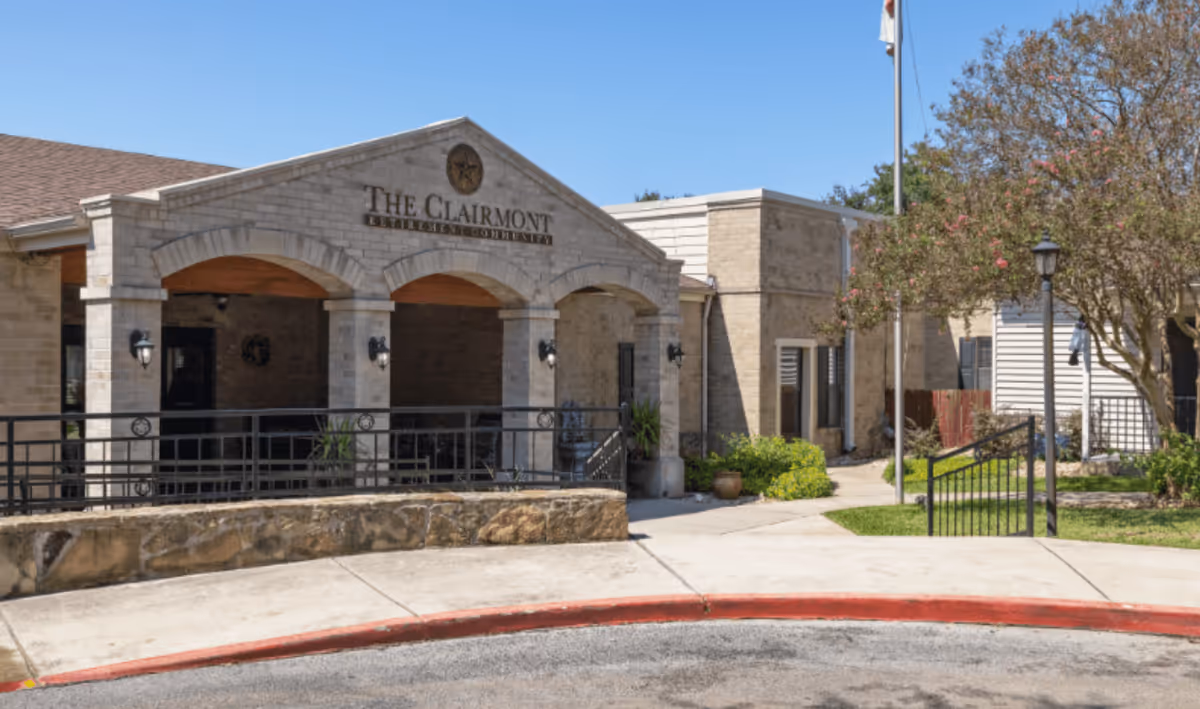 Stone-front entrance of The Clairmont Retirement Community with arched porch, railing, and a flagpole in front.