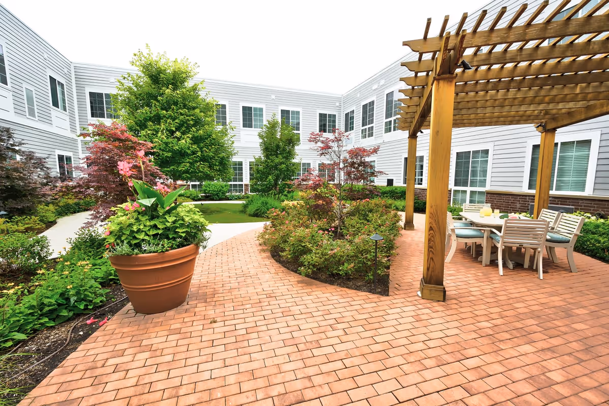 Outdoor courtyard area at Palos Heights Senior Living featuring a brick-paved patio with a wooden pergola covering a dining table and chairs. The courtyard is surrounded by well-maintained greenery, including trees, shrubs, and a large potted plant with pink flowers. The building with multiple windows encloses the courtyard.