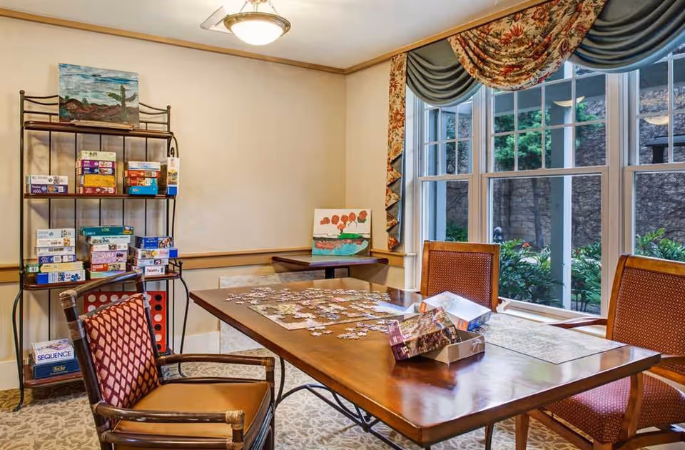 A cozy activity room with a wooden table in the center covered with a partially completed jigsaw puzzle and puzzle boxes. Three cushioned chairs surround the table. A metal shelf against the wall holds various board games and puzzles. Large windows with floral and green drapes let in natural light and show greenery outside. Two paintings are displayed on the shelf and table against the wall.