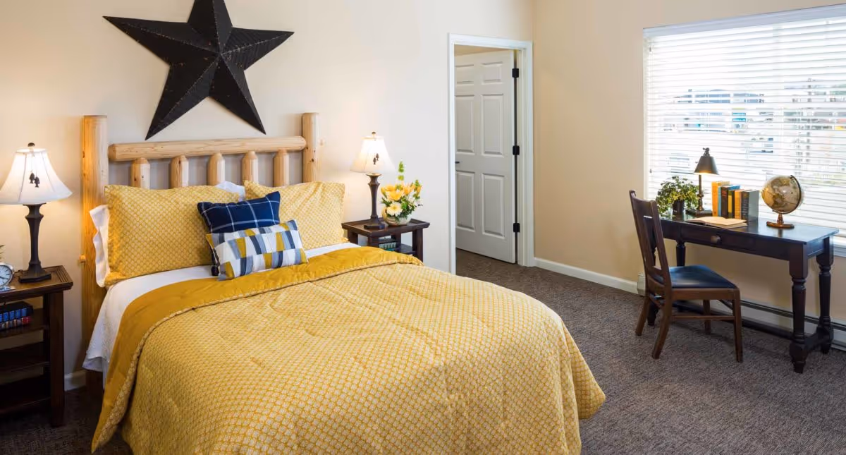 A cozy bedroom with a wooden bed frame and yellow bedding, two bedside tables with lamps, a decorative black star on the wall above the bed, and a dark wooden desk with a chair near a window with blinds. The room has beige walls and carpeted floor.