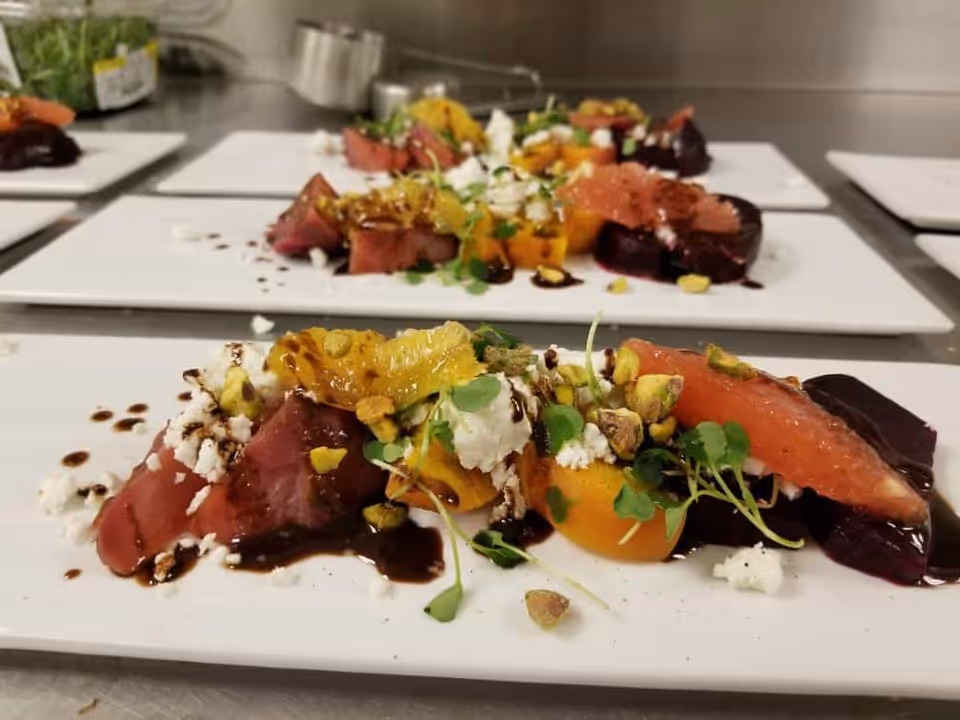 Plated gourmet salads with beets, citrus segments, goat cheese, microgreens and pistachios on white rectangular plates on a stainless prep counter.