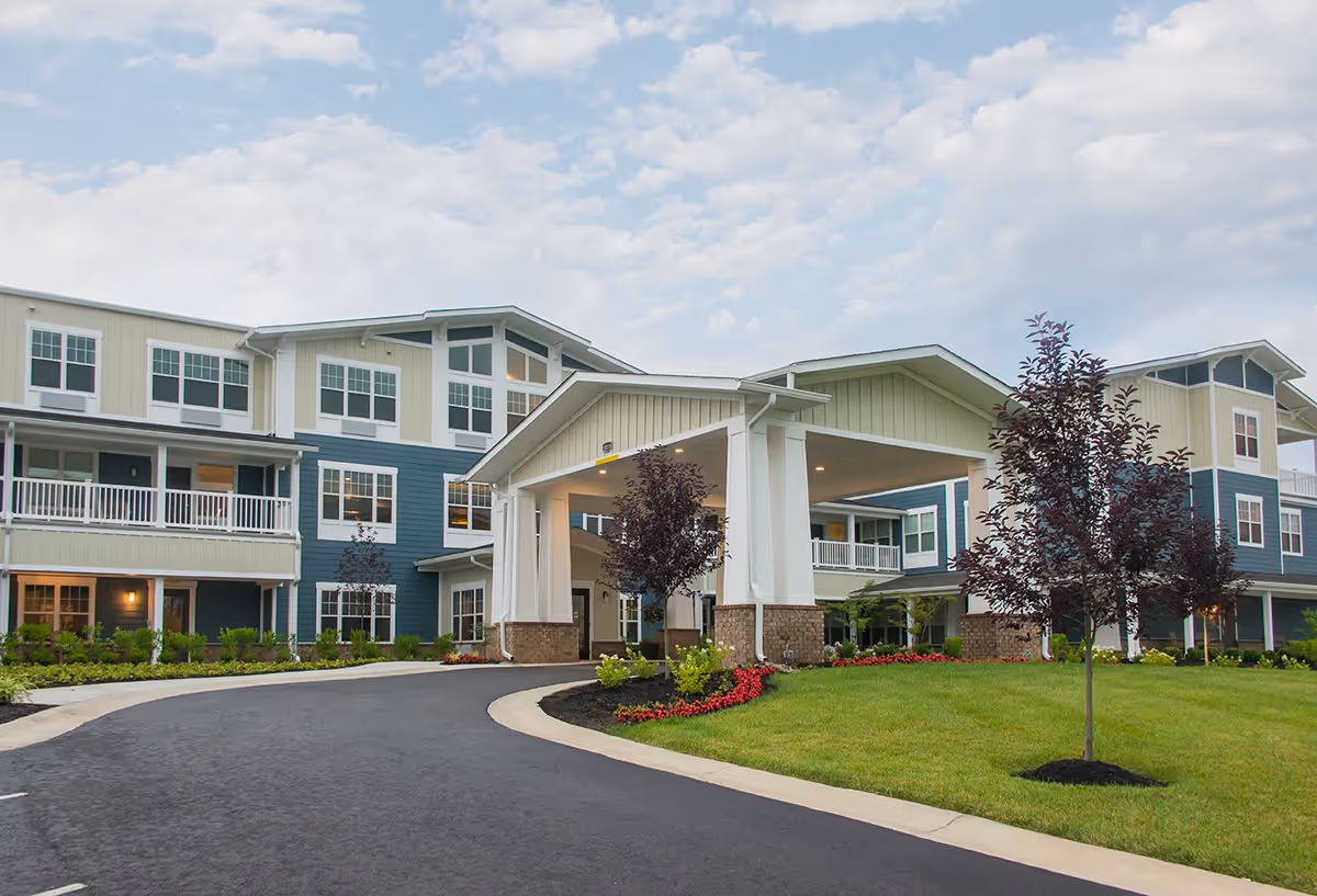 Exterior view of a multi-story senior living facility with a covered entrance, landscaped lawn, and young trees under a partly cloudy sky.