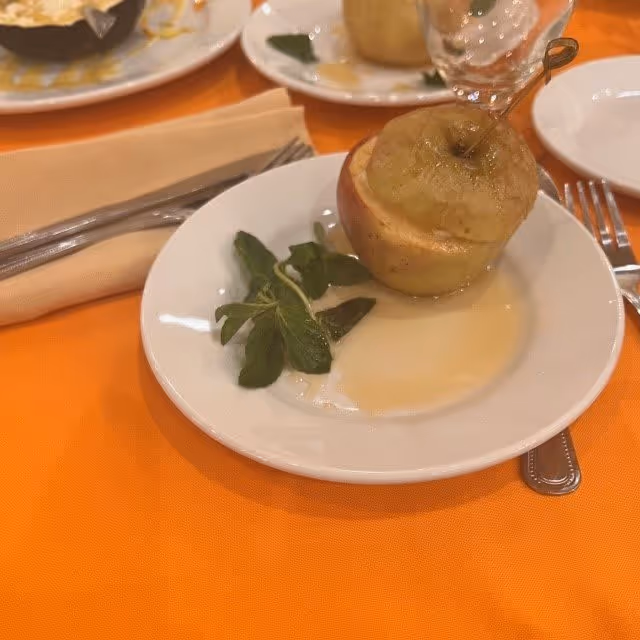 A white plate with a baked apple dessert garnished with a sprig of mint on an orange tablecloth. There is a beige napkin with a fork and knife beside the plate, and another plate with a similar dessert and a glass in the background.