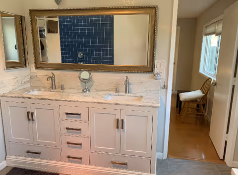 A bathroom vanity with a white marble countertop featuring two sinks and silver faucets. Above the vanity is a large rectangular mirror with a gold frame. To the left, there is a smaller mirror on the wall. In the reflection of the large mirror, blue tiled shower walls and a showerhead are visible. To the right, an open doorway leads to a room with wooden flooring, a window with blinds, and a wooden chair with a cushion.